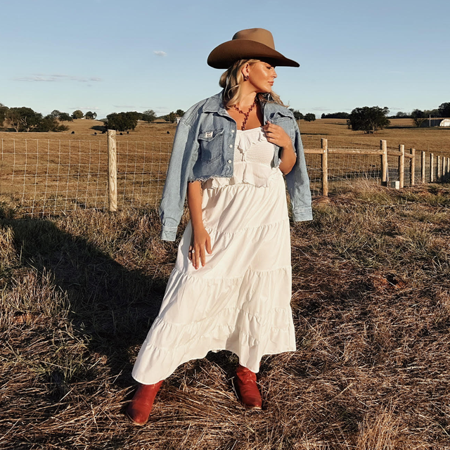 A woman standing in a field wearing a cowboy hat, a white dress and Justin western boots. 